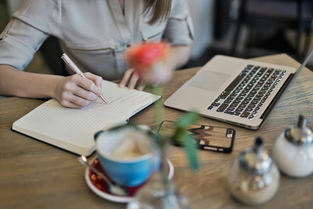Getboostify woman writing in a notebook with a laptop and coffee cup on a desk. Ideal for workspace inspiration.